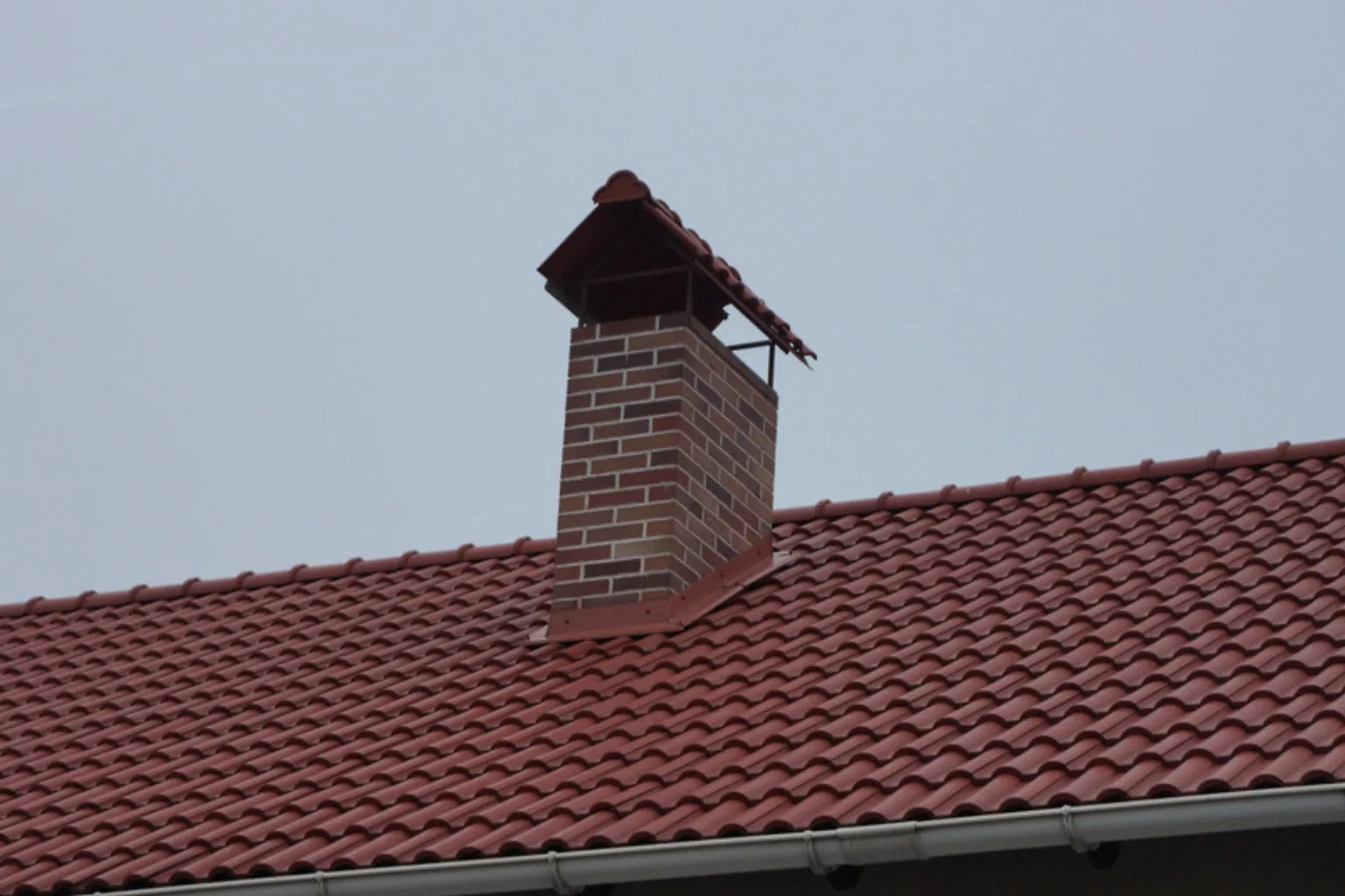 Murstensskorsten med hætte på et rødt tegltag mod en grå himmel